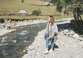 brunetta giovane donna viaggiatore nel poncho vicino il montagna fiume, nazione lato foto