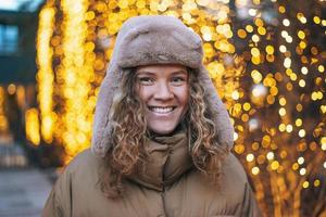 ritratto di giovane sorridente donna con Riccio capelli nel pelliccia cappello nel il inverno strada decorato con luci foto