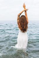 giovane spensierato donna con lungo capelli nel bianca vestito godendo vita su mare spiaggia, persone a partire dal dietro a foto