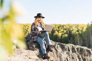 giovane bellissimo donna con Riccio capelli nel provato cappello e plaid camicia utilizzando il computer portatile su Visualizza sfondo di montagne, escursioni a piedi su il autunno natura, concetto di libero professionista foto