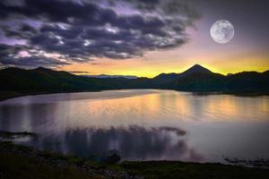 paesaggio lago fiume natura con pieno Luna tramonto giallo e viola cielo buio nuvole in movimento su silhouette montagna foto