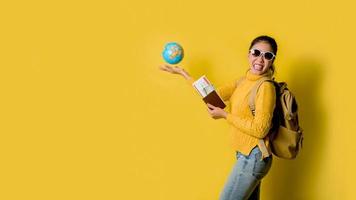 donna viaggiatore con valigia, Tenere palla globo nel il mano con passaporto e biglietto su giallo sfondo. ritratto di sorridente contento ragazza con spazio copia il testo. viaggio zaino foto
