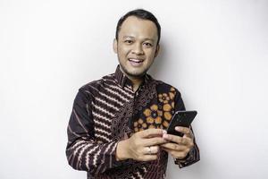 un' ritratto di un' contento asiatico uomo indossare batik camicia e Tenere il suo Telefono, isolato di bianca sfondo foto