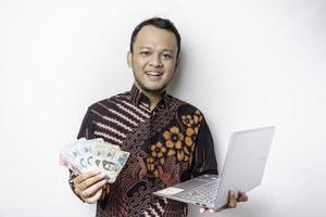 un' contento giovane asiatico uomo è indossare batik camicia, Tenere un' il computer portatile e i soldi nel indonesiano rupia isolato di bianca sfondo foto