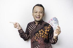 un' contento giovane asiatico uomo è indossare batik camicia, puntamento a il copia spazio accanto lui mentre Tenere i soldi nel indonesiano rupia isolato di bianca sfondo foto