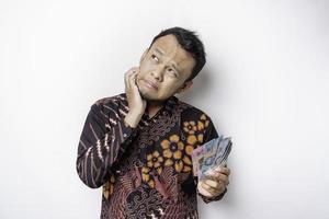 un' stressato asiatico uomo indossare batik camicia e Tenere i soldi nel indonesiano rupia isolato di bianca sfondo sembra depresso foto