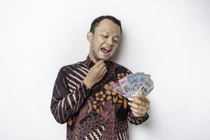 un' riflessivo giovane asiatico uomo è indossare batik camicia e Tenere denaro contante i soldi nel indonesiano rupia isolato di bianca sfondo foto