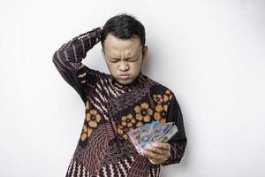 un' stressato asiatico uomo indossare batik camicia e Tenere i soldi nel indonesiano rupia isolato di bianca sfondo sembra depresso foto