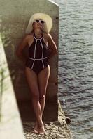 ritratto di un' donna nel un' costume da bagno, cappello e occhiali da sole nel estate su il Riva del fiume di un' calcestruzzo parete foto