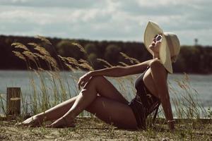 donna nel un' costume da bagno, cappello e occhiali da sole prende il sole nel estate su il Riva del fiume tra il erba foto