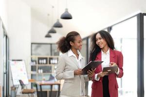 sorridente diverso colleghi donne d'affari Lavorando su il computer portatile insieme, guardare a schermo, In piedi a scrivania nel ufficio, dipendenti discutere progetto strategia, condivisione idee foto