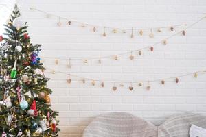 Natale albero nel bianca interno di un' Casa con in stile loft mattone muri con ghirlande di bicchiere giocattoli su un' corda. raggiante Fata luci decorazione di il studio camera. accogliente casa nel Natale e nuovo anno foto