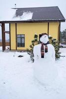 divertente pupazzo di neve nel un' cappello e sciarpa su il sfondo di un' giallo Casa nel il cortile. inverno, inverno intrattenimento, nevicata foto