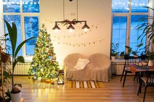 Natale albero nel bianca interno di un' Casa con in stile loft mattone muri con ghirlande di bicchiere giocattoli su un' corda. raggiante Fata luci decorazione di il studio camera. accogliente casa nel Natale e nuovo anno foto