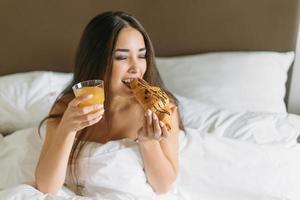 bellissimo giovane asiatico donna con lungo capelli nel avendo prima colazione brioche e arancia succo nel letto di Hotel camera foto