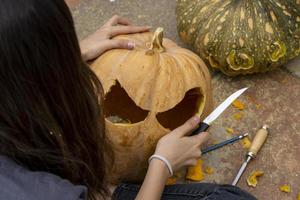 femmina intaglio grande arancia zucca per Halloween mentre seduta a di legno tavolo a casa foto