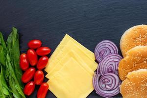 preparazione di un' Hamburger, tutti ingredienti formaggio, insalata, pomodoro, cipolla, ciambella menzogna su un' nero pietra sfondo. foto