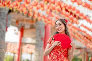 bellissimo asiatico donna sorridente felicemente Tenere ang pao, rosso buste indossare cheongsam guardare fiducioso nel Cinese buddista tempio. celebrare Cinese lunare nuovo anno, festivo stagione vacanza foto