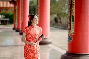 un' asiatico donna indossare tradizionale cheongsam qipao vestito Tenere fan e Sorridi mentre visitare il Cinese buddista tempio. Cinese nuovo anno concetto foto