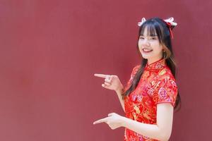 carino asiatico ragazza con lungo capelli chi indossa un' rosso cheongsam vestito nel Cinese nuovo anno tema mentre lei Spettacoli sua mano per presente qualcosa su un' rosso sfondo. foto