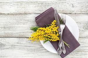 festivo tavolo ambientazione piatto con tovagliolo forchetta e coltello e mimosa fiore decorazione su bianca di legno sfondo foto