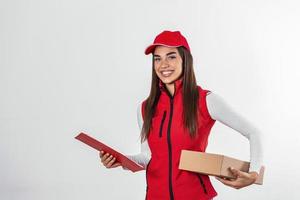 consegna persona consegna pacchi Tenere appunti e pacchetto sorridente contento nel rosso uniforme. bellissimo giovane donna professionale Corriere isolato su bianca sfondo foto