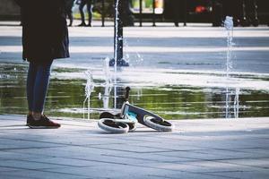 mamma con del bambino scooter dire bugie su marciapiede vicino Fontana enorme acqua pozzanghera con verde alberi riflessione, solitario genitore in attesa per ragazzo foto