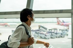 un asiatico uomo sta Tenere un' zaino nel il aeroporto finestra mentre in attesa per un' volo a il imbarco cancello prima partenza. foto