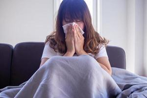 lungo dai capelli donna seduta su il divano è sofferenza a partire dal influenza, tosse e starnutire. seduta nel un' coperta perché di alto febbre e copertina loro naso con fazzoletto di carta carta perché starnuti tutti il volta. foto