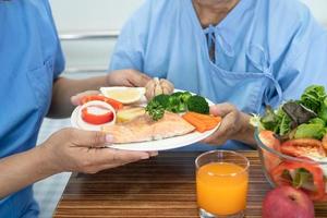 paziente asiatico anziano o anziano donna che mangia bistecca di salmone colazione con cibo sano vegetale mentre è seduto e affamato sul letto in ospedale. foto