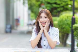 ritratto di un asiatico tailandese ragazza alunno nel un' uniforme e inserire fermo è seduta sorridente felicemente e con fiducia mentre toccante il tuo viso con il tuo mani nel Università foto