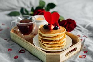 fatti in casa Pancakes con frutti di bosco, tazza di tè e fiori e regalo scatola. prima colazione o brunch per San Valentino giorno. foto