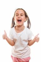 contento bambino poco ragazza mostrando pollici su gesto nel un' bianca t camicia isolato su bianca sfondo foto