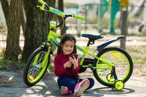 bambino in viaggio bicicletta nel estate parco. ciclista poco ragazza orologio su mobile Telefono. ragazzo conta pulse dopo sport formazione e è guardare per modo per navigatore. foto