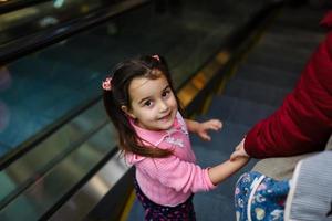 carino poco bambino in piedi su in movimento scala di il mano con mamma foto