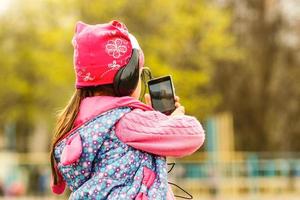 poco ragazza scolaretta. estate nel natura. nel mani Tenere un' smartphone ascoltando per musica su cuffia. prendere un' immagine su Telefono, parlando su il video chiamata. emozione sorrisi felicemente. foto