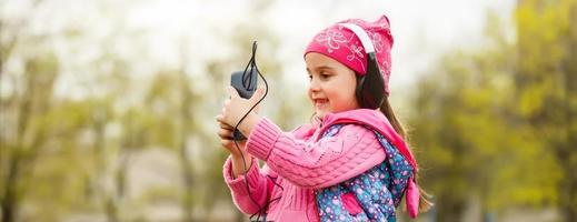 lato Visualizza di contento giovane ragazza bambino indossare colorato cuffia, ascoltando per musica su il smartphone all'aperto nel estate. foto