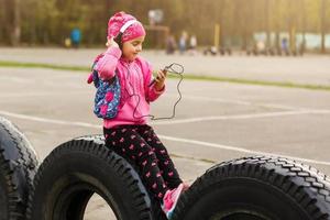 poco ragazza nel un' gonna seduta su un' parco panchina ascoltando per musica e guardare a mobile Telefono foto