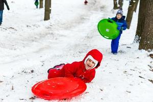 bella sorridente poco ragazza nel sua sciare completo da uomo scorrevole giù un' piccolo neve coperto collina con sua slitta foto