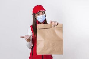 consegna donna dipendente nel rosso berretto maglietta uniforme maschera guanto hold mestiere carta pacchetto con cibo isolato su bianca sfondo studio servizio quarantena pandemia coronavirus virus 2019-ncov concetto foto