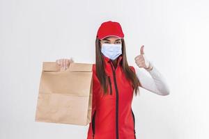 consegna donna dipendente nel rosso berretto maglietta uniforme maschera guanto hold mestiere carta pacchetto con cibo isolato su bianca sfondo studio servizio quarantena pandemia coronavirus virus 2019-ncov concetto foto