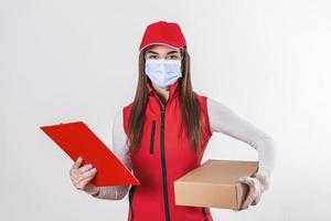 consegna persona consegna pacchi Tenere appunti e pacchetto sorridente contento nel rosso uniforme. bellissimo giovane donna indossare medico maschera e guanti professionale Corriere isolato su bianca sfondo foto