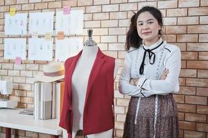 asiatico adolescente femmina moda progettista sembra a telecamera e sorrisi nel studio, braccia attraversato, Lavorando con misurare nastro e cucire per vestito design collezione, giovane boutique sarto pmi avviare imprenditore. foto