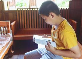 un' bianca asiatico ragazzo con un' fossetta su il suo guancia felicemente si siede giù e legge un' libro per revisione il Lezioni a casa. foto