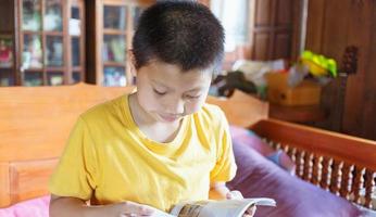 un' bianca asiatico ragazzo con un' fossetta su il suo guancia felicemente si siede giù e legge un' libro per revisione il Lezioni a casa. foto