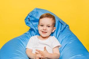 sorridente poco ragazzo seduta nel un' blu sedia su un' luminosa giallo sfondo foto