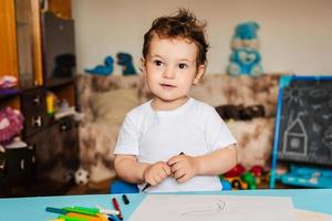 un' piccolo ragazzo disegna su lenzuola di carta dire bugie su il tavolo con colorato matite foto