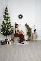 ritratto di bella giovane ragazza accogliente sedersi giù Tenere Natale regalo, sorridente indossare rosso toga nel decorato Natale vivente camera in casa foto