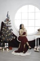ritratto di bella giovane ragazza accogliente sedersi giù e potabile vino, indossare rosso toga nel decorato Natale vivente camera in casa foto
