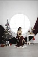 ritratto di bella giovane ragazza accogliente sedersi fuori uso, sorridente indossare rosso toga nel decorato Natale vivente camera in casa foto